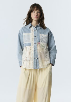 Jeune femme aux cheveux châtain moyen porte une chemise en denim bleu avec des patchs en dentelle crème et un pantalon ample crème, debout devant un fond clair.