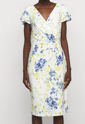 Žena v bílých šatech po kolena s modrým a žlutým květinovým vzorem a krátkými volánovými rukávy, stojící před jednoduchým pozadím.