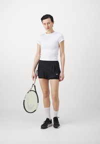 Weißes Kurzarmhemd, schwarze plissierte Shorts, weiße knielange Socken und schwarze Sneaker. Hält einen Tennisschläger mit gemustertem Griff.