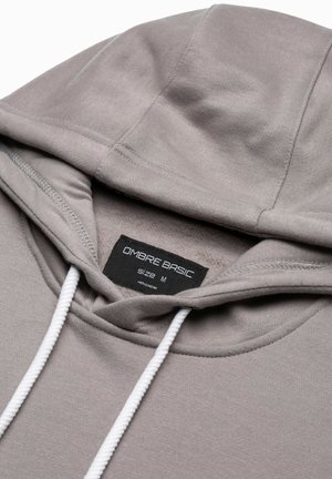 Sudadera gris claro con cordones, tela suave, bolsillo canguro y etiqueta que dice "OMBRE BASIC, Talla M." Detalles de costura simples.