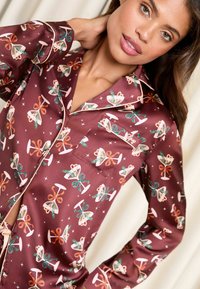 Camicia da pigiama color bordeaux con motivo floreale a fiocco, texture in raso, colletto a revers, taschino singolo sul petto e dettagli in piping lungo i bordi.