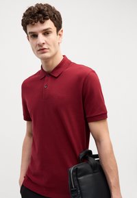 Burgundi polo marškiniai su trumpomis rankovėmis, trijų mygtukų užsegimas ir iškilmingas logotipas; laikomi su juodu odiniu krepšiu, turinčiu užtrauktuką.