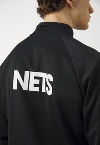 Giacca sportiva nera con colletto alto, realizzata in tessuto liscio, con grande stampa bianca "NETS" sul retro.