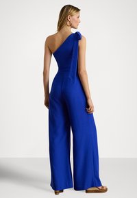 Modrý overal na jedno rameno so stuhou, širokými nohavicami a priliehavým živôtikom, vyrobený z hladkej látky s matným povrchom.