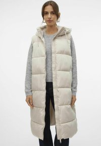 Gilet rembourré beige sans manches avec une capuche, doté d'une fermeture éclair sur le devant et de poches latérales, porté sur un pull gris et un jean foncé.