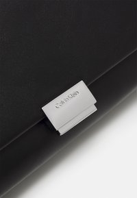Čierna kožená kabelka s hladkou textúrou, vybavená striebornou kovovou sponou s vyrazeným nápisom "Calvin Klein." Jednoduchý a obdĺžnikový dizajn.