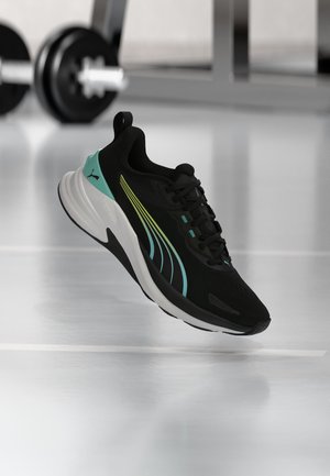 Chaussure de sport noire et turquoise avec des rayures jaunes et bleues flottant au-dessus du sol d’une salle de sport avec du matériel d’haltérophilie en arrière-plan.