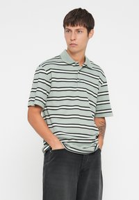 Striped polo shirt in muted green with black and white stripes. Short sleeves, classic collar, and a three-button placket. Worn with black pants.