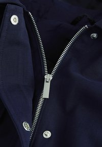 Mornarskoplavi sako sa srebrnim metalnim patentnim zatvaračem djelomično otvoren, prikazujući detalje tkanine i dva dugmeta na obje strane.