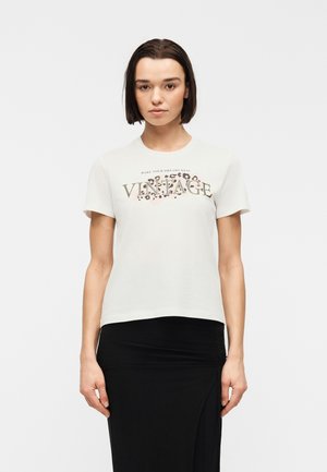 Sirge tumedate juustega naine, kes kannab valget "VINTAGE"-trükiga t-särki ja musta seelikut, seisab ühtlasel heledal taustal.