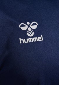 Mørkeblå stof præsenterer et fremtrædende hvidt Hummel-logo, der viser et abstrakt bierdesign og stiliseret tekst under det. Tekstureret overfladedetalje synlig.