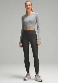 Grauer, langärmeliger Crop-Top kombiniert mit hoch taillierten schwarzen Leggings. Das Modell trägt hellgraue Turnschuhe und steht vor einem neutralen Hintergrund.