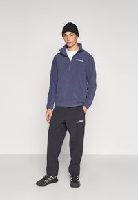 Granatowy polar z zamkiem przy szyi, zestawiony z czarnymi spodniami z akcentami logo. Model ma na sobie czarną czapkę typu beanie i buty outdoorowe.