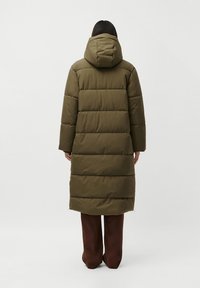 Person facing away wearing long olive green padded hooded coat and dark brown wide-leg pants against plain background.