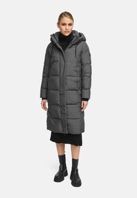 Cappotto imbottito grigio con cappuccio, caratterizzato da un design lungo, texture a righe verticali e tasche laterali, indossato sopra un vestito di maglia nero e stivali.