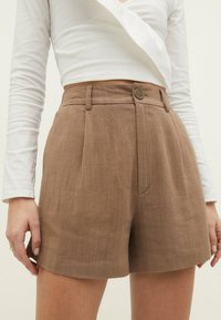 Bruine linnen short met een slanke pasvorm, voorzien van een knoopsluiting, plooien en zijzakken. Gecombineerd met een wit top met lange mouwen.