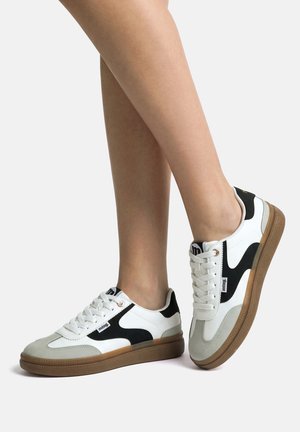 Persona con zapatillas blancas con detalles negros y suelas de goma, de pie con un pie ligeramente levantado contra un fondo liso.