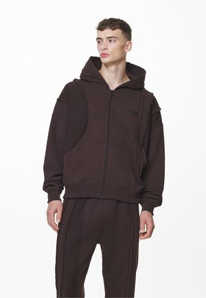 Brun zip-up hoodie med en frontlomme, raglanærmer og en stor hætte, parret med matchende bukser med kontrasterende sømme.