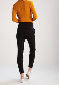 Kvinna iförd en senapsgul ribbad tröja, svarta åtsittande byxor och silverfärgade slip-on skor, stående med ryggen mot kameran.