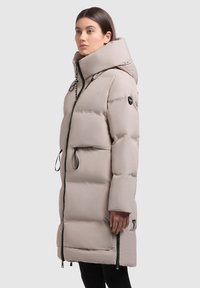 Világos bézs színű puffertakaró, magas gallérral, cipzáras zárással, húzózsinórokkal és oldalsó cipzáras zsebekkel. A ujjon logó felvarrás található.