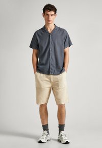 Short-sleeve, dark gray button-up shirt with a relaxed collar. Beige shorts with multiple pockets. Gray socks and white sneakers complete the look.