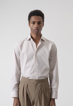 Joven con cabello corto y rizado, vistiendo una camisa clara de rayas abotonada y pantalones beige plisados de cintura alta, de pie contra un fondo liso.