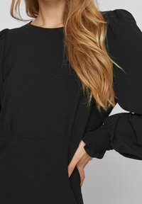 Robe noire avec de longues manches bouffantes et des poignets smockés. Le tissu a une texture subtile et il y a des poches sur les côtés.