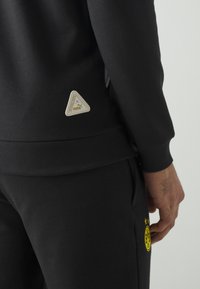 Svart atletisk sweatshirt med ribbade muddarmar, med en triangelformad beige logotypapplikation på baksidan och subtila grå detaljer på sidorna.