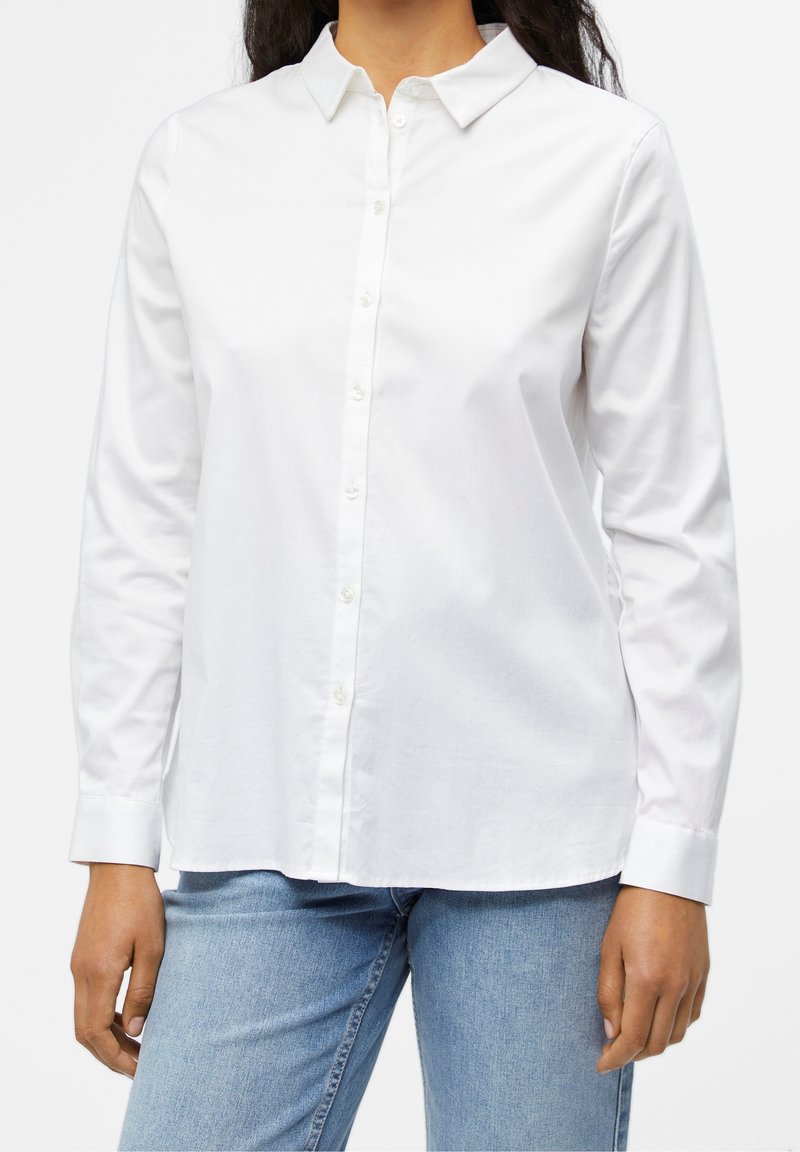 Camisa blanca de algodón con cuello extendido, mangas largas y un ajuste relajado, combinada con vaqueros de color azul claro. Diseño liso.