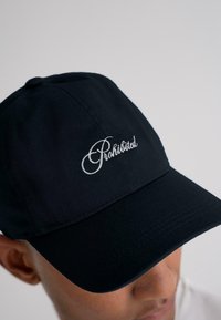 Persona che indossa un cappellino da baseball nero ricamato con la parola "Proibito" in carattere corsivo bianco sulla parte anteriore.