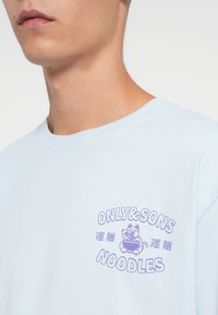 Lichtblauw shirt met een paarse grafiek van een kat die noedels eet en de tekst "ONLY & SONS NOODLES" met Aziatische karakters.