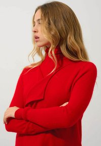 Roter Rollkragenpullover aus weichem Strickmaterial, mit einem drapierten Halsausschnitt und gerippten Bündchen. Das Design ist figurbetont und hat lange Ärmel.