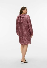 Glitrende pink kjole med lange ærmer, en sløjfe bagpå og løs pasform. Har pailletter, der dækker hele stoffet. Sorte mules med hæl er båret.