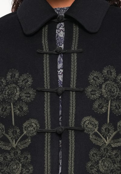 Μαύρο παλτό από μαλλί με γιακά, διαθέτει πράσινη λουλουδάτη κεντητική και διακοσμητική ραφή, κλείνει με κουμπιά-χάρακες. Ορατό μοτίβο από εσωτερική στρώση.