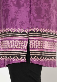 Lila Tunika aus einem strukturierten Stoff mit einem lebhaften Muster aus pinkfarbenen, schwarzen und weißen geometrischen Designs. Seitenschlitze verbessern die Bewegungsfreiheit.