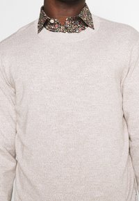 Pull beige clair en maille avec une finition texturée, superposé à une chemise noire à col à motif floral avec des boutons blancs.