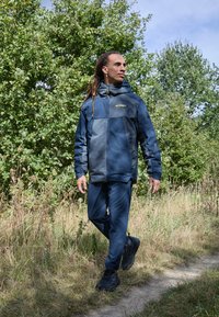 Blau- und graue Outdoorjacke mit Kapuze, passende Hose und schwarze Schuhe. Merkmale sind Reißverschlüsse und ein Logo auf der Brust.