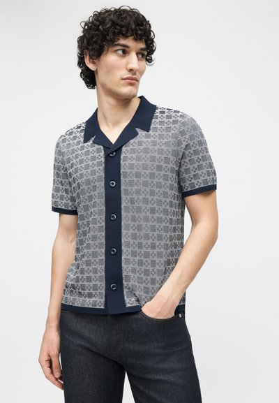 Jeune homme aux cheveux bouclés portant une chemise à manches courtes à motifs et un jean foncé, debout avec une main dans la poche.