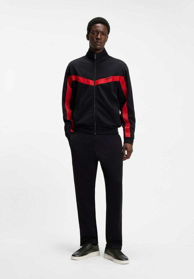 Jeune homme debout portant un survêtement noir avec des bandes rouges et des baskets noires sur fond blanc.