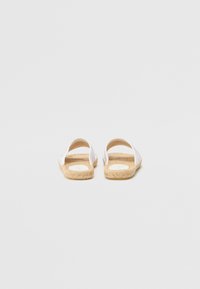 White woven fabric sandals with a natural jute sole, open back, and crisscross strap design. Flat sole enhances casual style.