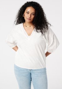 Weißes V-Ausschnitt-Top mit strukturierten Puffärmeln und Spitzendetails am Ausschnitt, kombiniert mit hellblauen, figurbetonten Jeans.