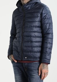 Granatowa pikowana kurtka z zamkiem błyskawicznym z przodu i kapturem. Charakteryzuje się poziomym przeszyciem oraz małym logo na dole. Noszona z dżinsami.