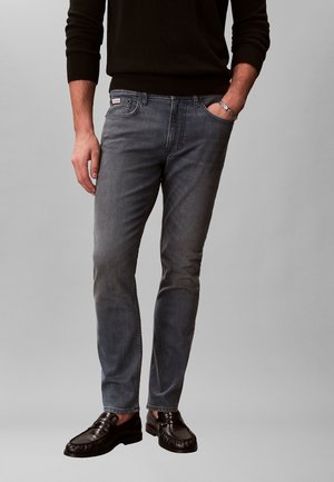 Mann trägt ausgewaschene graue Slim-Fit-Jeans, schwarze Loafers, schwarzes Langarmoberteil, mit der Hand in der Tasche und silbernem Armband am Handgelenk.