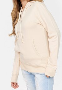 Felpa beige con tasca frontale, cappuccio con cordino, polsini a coste e vestibilità rilassata. Tessuto morbido con una superficie liscia. Indossata con jeans azzurri chiari.
