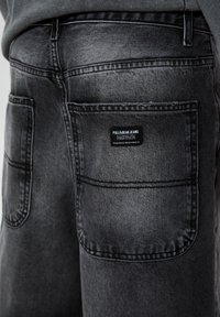 Jean en denim noir à l'aspect délavé, avec deux poches arrière et une étiquette portant le nom de la marque sur la poche droite.