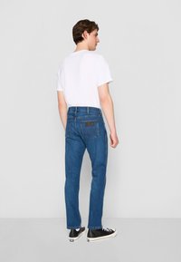 Vaqueros de mezclilla azul con un corte recto, que cuentan con una etiqueta de cuero marrón en la parte trasera, y una camiseta blanca. Zapatillas negras completan el look.