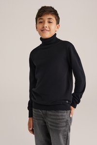 Melnas turtleneck džemperis, izgatavots no mīkstas auduma ar ribotām apkakli un apakšmalu. Valkāts ar pelēkiem džinsiem, kas piedāvā nelielu logotipa detaļu.