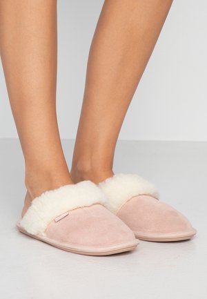 Füße, die hellpinkfarbene Slip-On-Hausschuhe mit weißem, flauschigem Innenfutter tragen, stehen auf einem glatten grauen Boden vor einem schlichten grauen Hintergrund.