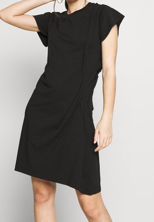 Kobieta w czarnej sukience z krótkim rękawem, z subtelnymi detalami szwów, stojąca na tle jednolitego jasnego tła.