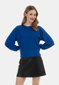 Blaues langärmliges Oberteil mit rundem Halsausschnitt und Puffärmeln; kombiniert mit einem schwarzen Leder-Minirock. Glatte Textur und taillierter Schnitt.